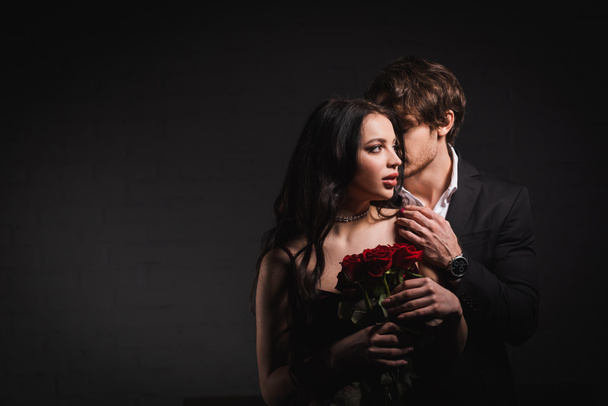 giovane uomo elegante abbracciare sensuale donna bruna che tiene rose rosse su sfondo scuro - Foto, immagini