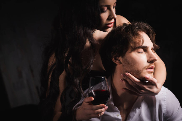 vášnivá brunetka žena drží sklenici červeného vína při objímání krku muže v bílé košili na tmavém pozadí - Fotografie, Obrázek