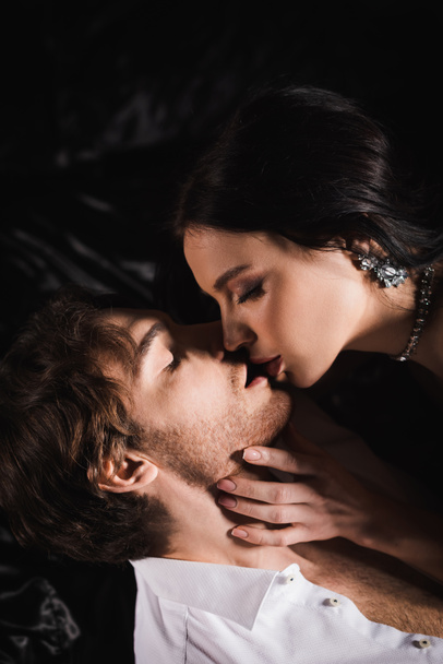 sensual woman in jewelry kissing man in white shirt on black bedding at night - Foto, afbeelding