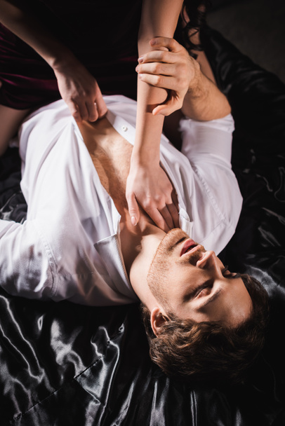 high angle view of woman undressing man in white shirt lying with closed eyes on black satin bedding - 写真・画像