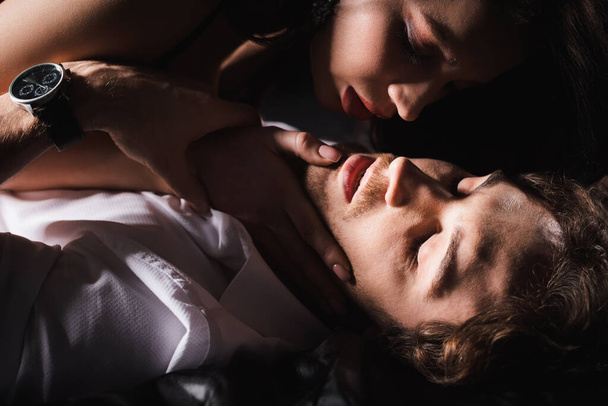 страстная женщина трогает лицо мужчины, лежащего с закрытыми глазами в темноте - Фото, изображение