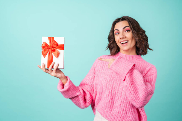 Acogedor retrato de una mujer joven en un suéter de punto rosa y con maquillaje brillante sobre un fondo turquesa con una caja de regalo con un lazo rojo - Foto, Imagen