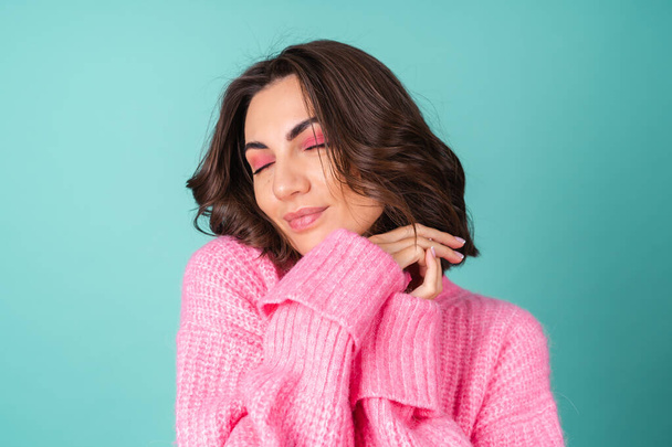 Acogedor retrato de una mujer joven en un suéter de punto rosa y con maquillaje brillante sobre un fondo turquesa - Foto, imagen
