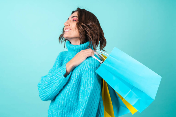 Cozy portrait of a young woman in a knitted blue sweater and bright pink makeup on a turquoise background holding shopping bags - Foto, Imagem