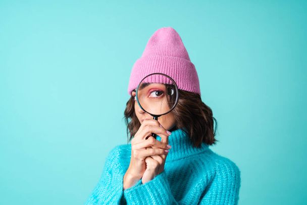 Portrait confortable d'une jeune femme dans un pull bleu tricoté et un chapeau rose avec un maquillage lumineux tenant une loupe, s'amuser, s'amuser - Photo, image