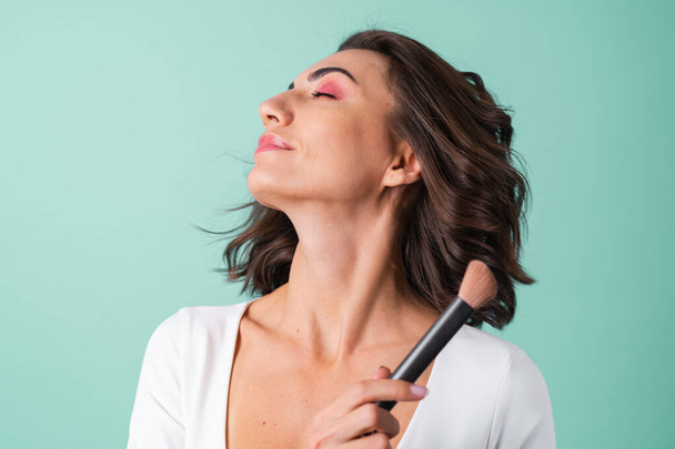 Young woman in a white evening dress on a light green background with bright pink makeup holding a blush brush - Фото, изображение