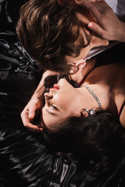 overhead view of blurred man near passionate woman in jewelry lying on black bedding with closed eyes - 写真・画像