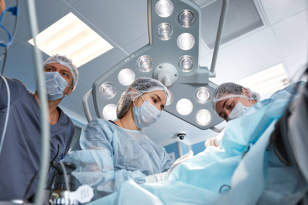 Konsantre cerrahi ekibi ameliyathanede bir hastayı ameliyat ediyor. Karmaşık makinelerle yıllarca eğitim almış iyi eğitimli bir anestezi uzmanı ameliyat boyunca hastayı takip eder. - Fotoğraf, Görsel