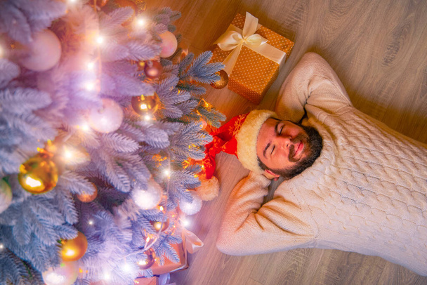 Christmas holidays festive happy season time middle adult man portrait take a relax laying pose on a floor in home under Christmas tree with garland lamps illumination - Zdjęcie, obraz