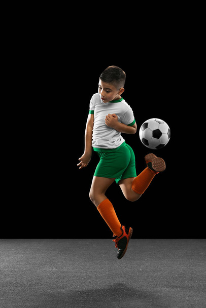 Full-length portrait of boy, child, football player in uniform training, kicking ball in a jump isolated over black background. Making backheel - Foto, Imagem