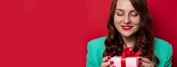 Beautiful young woman with expressing happy emotions and holding present box on a red wall background. - Фото, изображение