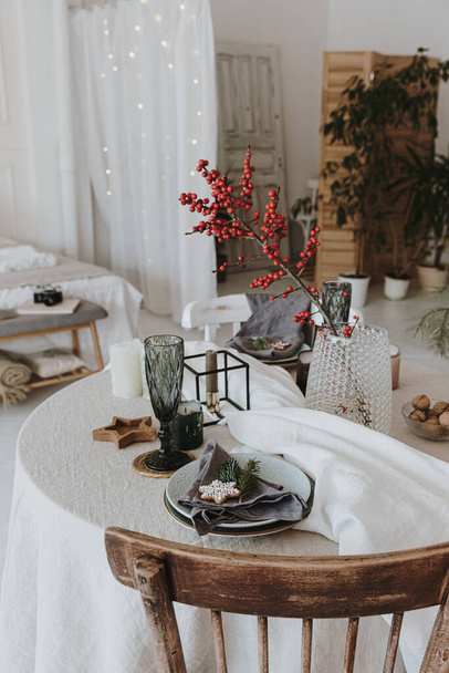 Elegant aesthetic Christmas dinner concept. Table with grey linen table cloth, red berries branch bouquet, gingerbread cookies, glass, plates. Christmas celebration evening - Photo, image
