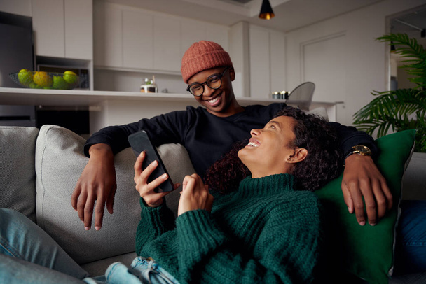 Multi-ethnic couple looking at each other, laughing about text on phone. On sofa in modern apartment - Foto, Imagen