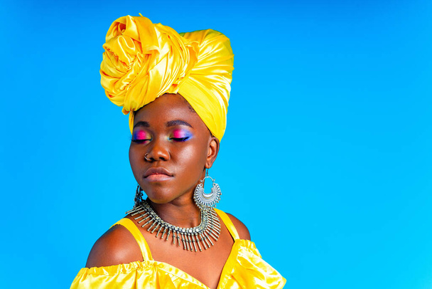 beautiful african woman in yellow silk turban on head and dress with silver neckless and earrings , nose piercing in blue studio background - Foto, Imagen