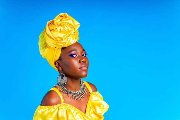 African woman with yellow silk turban and silver earrings and nose piercing in blue studio background - Фото, изображение
