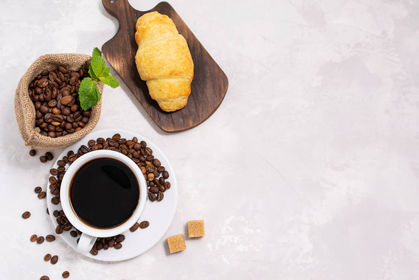 Fresh aromatic black coffee in a white ceramic cup on a gray-white textured background. Roasted coffee beans in a canvas bag. Croissant on a dark wooden board. Top view, space for text - Photo, Image