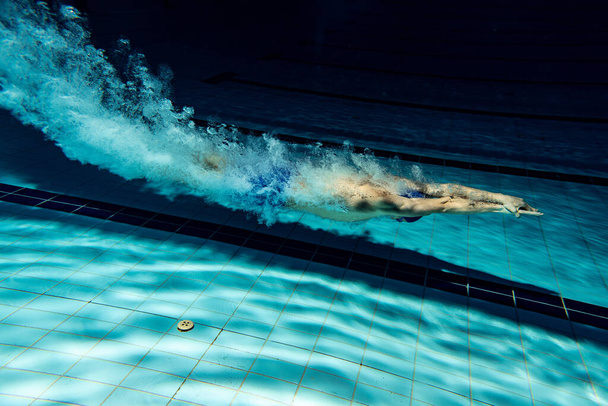 Underwater camera work. One male swimmer training at pool, indoors. Underwater view of swimming movements details. - Фото, изображение
