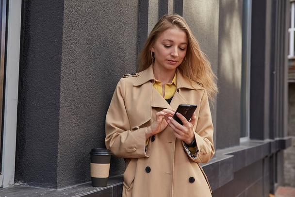caucasian woman in beige trench coat using smartphone outdoors on sunny day - Foto, Bild