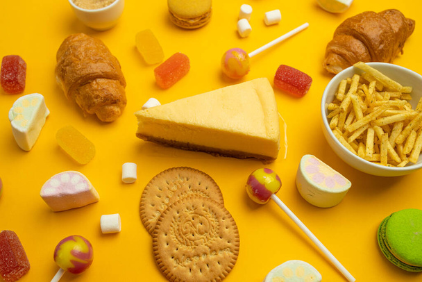 Alimentos não saudáveis em fundo colorido, vista superior. Croissant francês, doces em um pau, doces de geleia, marshmallows, biscoitos e macarons em um fundo brilhante amarelo - Foto, Imagem