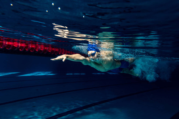 Close-up. Young male swimmer in swimming cap and goggles in motion and action during training at pool, indoors. Underwater imaging - Foto, Imagen