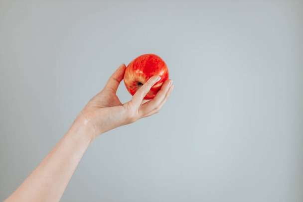 灰色の背景に新鮮な健康的な赤いリンゴを保持する女性の手。最小限のフルーツコンセプト. - 写真・画像