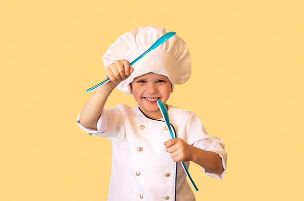 Bambino divertente in uniforme da chef bianco, che tiene le pinze per insalata tra le mani come un ninja. Allegro sfondo giallo. - Foto, immagini