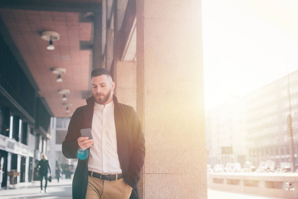 Young handsome caucasian bearded elegant man walking backlight outdoors city using smartphone browsing 5g internet - 写真・画像