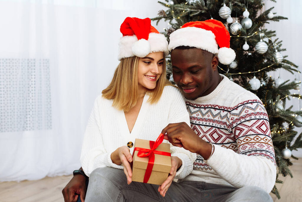 Fröhliche, gemütliche Momente in den Winterferien. Ein multinationales Paar öffnet ein Weihnachtsgeschenk. Ein liebevoller schwarzer Mann und eine weiße Frau sitzen neben dem Weihnachtsbaum - Foto, Bild