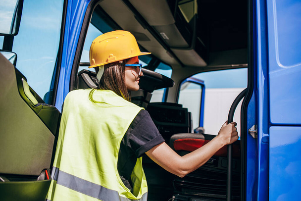 Portrait of beautiful young woman professional truck driver with protective yellow helmet sitting and driving a big truck. Inside of vehicle. People and industrial transportation concept. - Fotografie, Obrázek