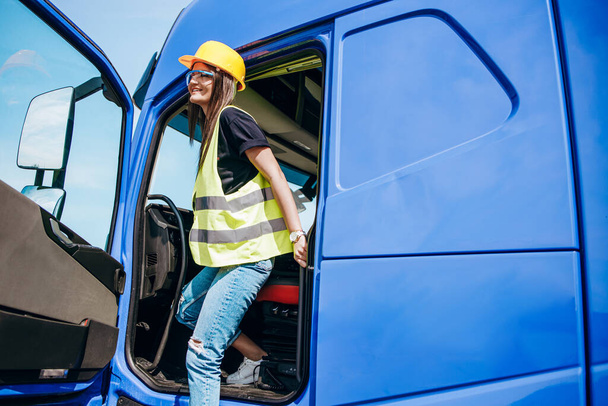 Portret van een mooie jonge vrouw professionele vrachtwagenchauffeur met beschermende gele helm zittend en rijdend in een grote vrachtwagen. Binnenkant van het voertuig. Concept van personen- en industrieel vervoer. - Foto, afbeelding
