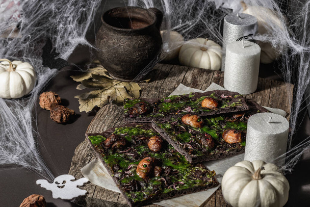 Halloween concept food. Flatbread or pizza with squid Ink, green cheese, mushrooms and purple onion. Bread of the Dead with festive decor, white pumpkin, candles and spider web. Dark stone background - Foto, afbeelding