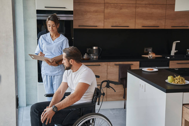 Beautiful doctor in uniform and male who using a wheelchair having conversation in the home kitchen - Zdjęcie, obraz