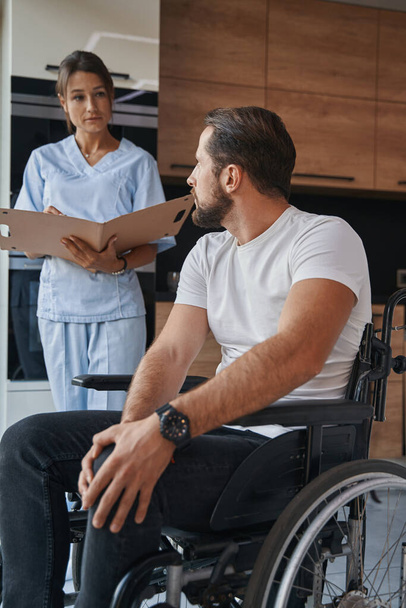 Adorable man who using a wheelchair talking with physiotherapist in the modern home apartments - Foto, Bild