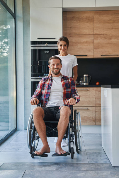 Adorable man who using wheelchair and charming female posing at the photo camera in the house - Φωτογραφία, εικόνα