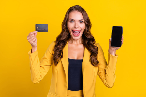 Photo of funny impressed young lady wear suit jacket holding bank card modern gadget empty space smiling isolated yellow color background - Fotoğraf, Görsel
