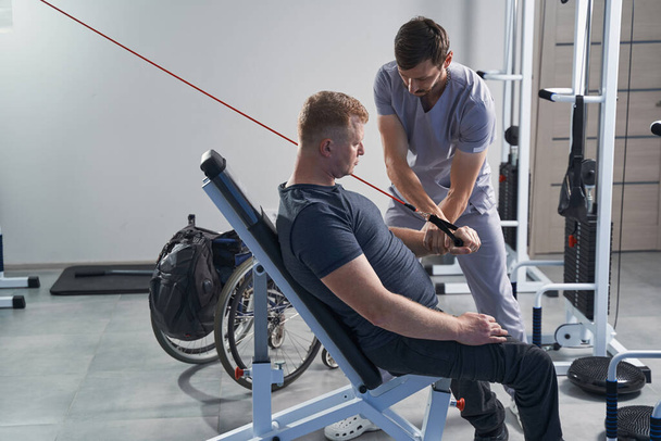 Man met een handicap riemen op gewichtheffen apparatuur met therapeut - Foto, afbeelding