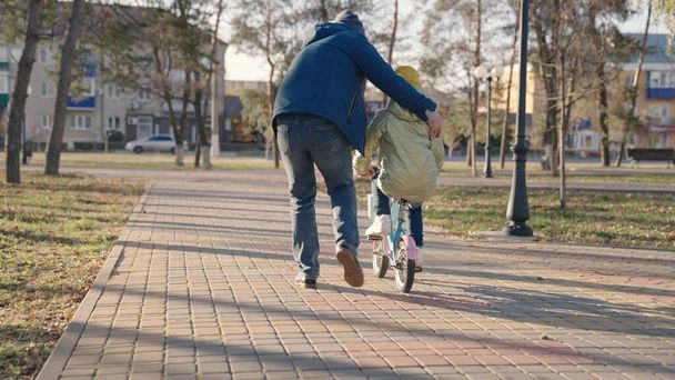 father teaches little kid to ride bike on road in city park, child spins pedals and wheels, happy family, dad helps the girl to ride daughter, play in autumn park, parent and baby have fun together - Photo, Image