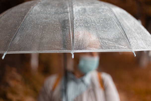 Vecchia donna in abiti luminosi sotto un ombrello trasparente e mano tesa cattura gocce d'acqua con contenuto in caso di maltempo nel parco della città - Foto, immagini