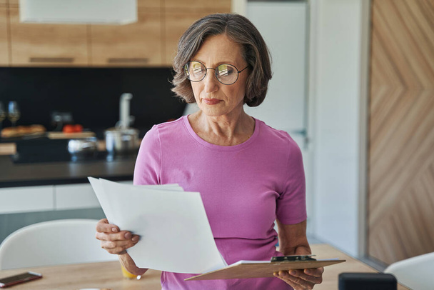 Senior lady looking through document in kitchen - Foto, imagen