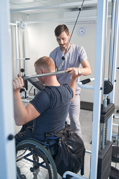 El médico ayuda al paciente en silla de ruedas en la máquina de peso en la sala de fisioterapia - Foto, imagen