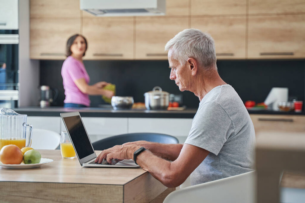 Elderly male in the kitchen sitting in front laptop - Foto, Bild