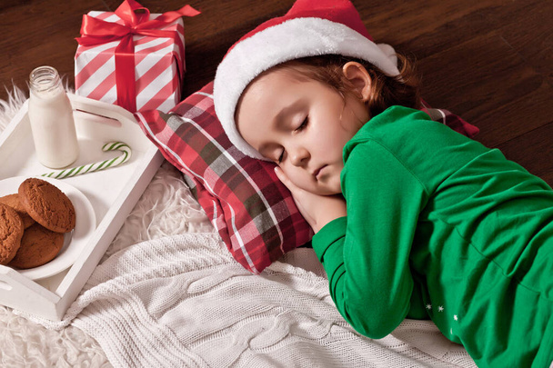 happy child girl in Santa cap waiting Santa and sleep near pine tree on Christmas eve. - Zdjęcie, obraz