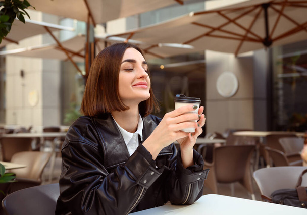 Delighted beautiful young woman with closed eyes of pleasure, cutely smiles enjoying coffee break, resting in outside cafe, holding a takeaway paper cup with hot drink. Freedom, freelancer, lifestyle. - Foto, afbeelding