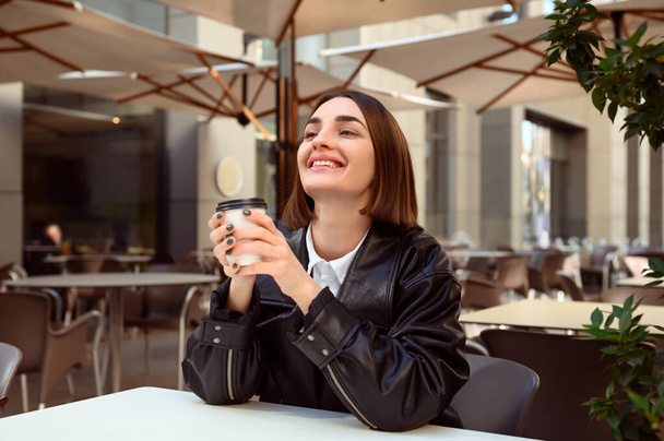 Beautiful young woman with closed eyes of pleasure, enjoying coffee break, resting in outside cafe, holding a takeaway paper cup with hot drink, smiling looking up. Freedom, freelancer, lifestyle. - Photo, image