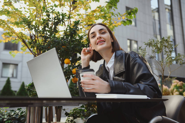 Smiling beautiful woman, freelancer, copywriter, developer enjoys coffee break in a terrace of cafeteria, works remotely on laptop, plans projects, looks at camera. Distant online business concept - Photo, image