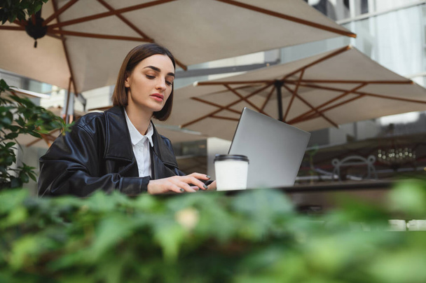 Beautiful concentrated woman, copywriter, freelancer working on laptop siting in summer terrace of a cafeteria. Distant remote online business concept - Photo, Image