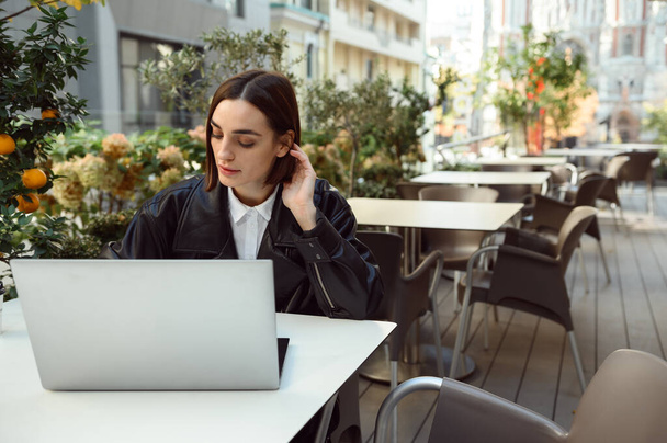 Online business and distant work concept. Beautiful concentrated freelance woman, young business person, copywriter, using laptop while sitting at table in a terrace of cafeteria and working remotely. - Photo, Image
