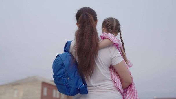 madre lleva a una pequeña hija con una mochila en sus brazos a la escuela, enseñando a un niño en la escuela primaria, una familia feliz, despedir a un niño con una bolsa de la escuela, adquirir conocimiento de la vida futura - Foto, Imagen