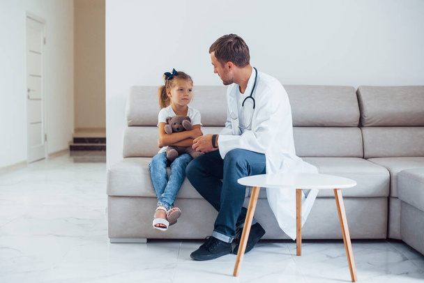 Male doctor in white uniform sits in the clinic with little girl. - Foto, Imagen