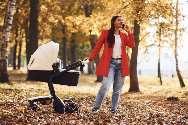 Talking by the phone. Mother in red coat have a walk with her kid in the pram in the park at autumn time. - Foto, afbeelding